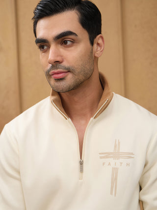 Men, looking to the side and wearing a cream-colored half-zip sweater with a cross and "FAITH" embroidered on the chest.