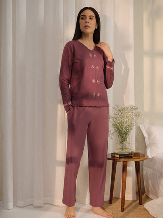Woman in plum pajamas with embroidered details stands posing next to a bedside table.
