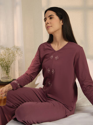 Woman sitting on a bed wearing a plum-colored pajama set with embroidered flowers.