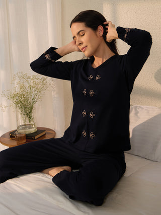 A woman in dark blue pajamas with floral accents sitting on a bed.