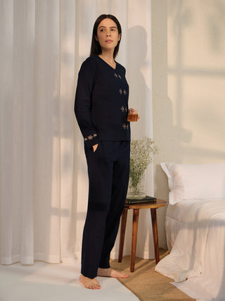 A woman in dark blue pajamas with floral accents, holding a drink, stands beside a bed.