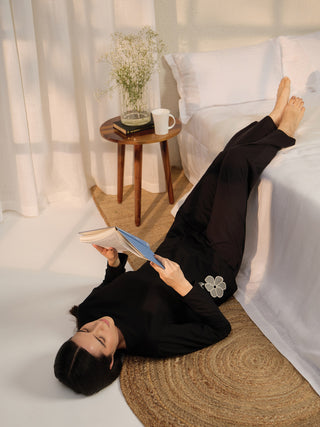 A woman in black sleepwear with a floral print is lying on the floor, reading a book.