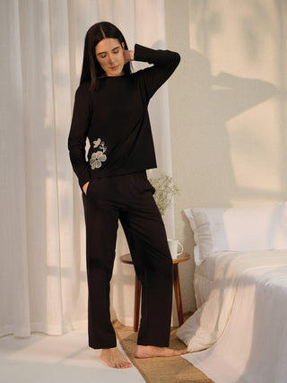 Woman in black long-sleeve pajama set with a floral print standing with one hand behind her head.