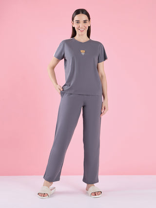 A woman stands with her left hand on her hip, wearing a grey short-sleeved t-shirt with a small teddy bear print and matching grey pants.