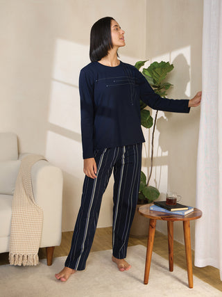 A woman in navy blue pajamas with a white pinstripe print stands by a curtain, holding it open.