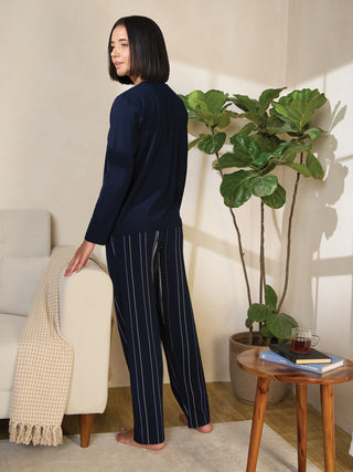 A woman in navy blue striped pajamas stands with her hand on a beige couch, a plant and side table with a drink and books are visible behind her.