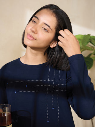 A woman in a dark blue long-sleeved sleep shirt with a delicate dashed-line and star print pattern, holding a glass of amber liquid in her left hand and touching her hair with her right.