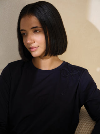 A woman wearing a navy blue long-sleeve sleep shirt with a delicate embroidered bow detail on the shoulder.