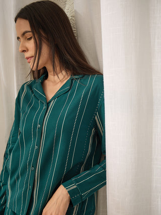 A woman leans against a white curtain, wearing a teal pajama top with vertical white and grey stripes.