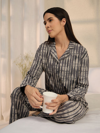A woman in grey and beige striped pajamas sits cross-legged on a bed holding a white mug.