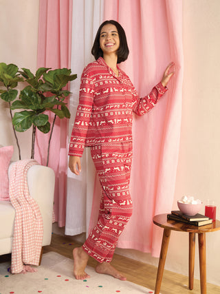 A woman in red holiday-print pajamas stands with her hand on a pink curtain, looking to the side and smiling.