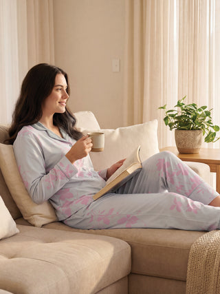 A woman in light blue floral print pajamas sits on a sofa, holding a mug and a book.