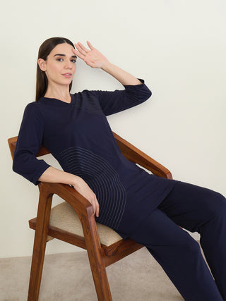 A woman sits on a wooden chair wearing a dark blue tunic with a curved line print and matching pants.