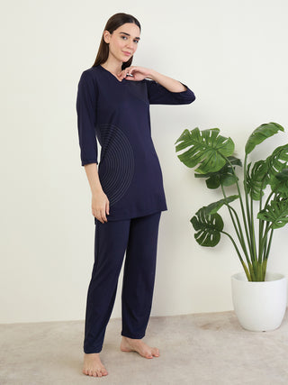 A woman stands in a navy blue pajama set with subtle concentric circle print, holding her hand to her neck.