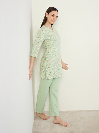 A woman in a light green printed tunic and matching pants leans against a white wall, her left hand touching the wall.
