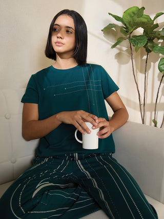 A woman in a teal t-shirt with a dashed line and star print and matching patterned pants, sitting on a couch holding a mug.