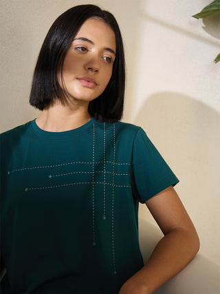A young woman with short black hair poses looking away from the camera wearing a dark teal t-shirt with a dashed line grid print and subtle star accents, contrasting the previous image with its different garment and print.