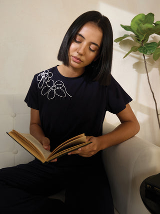 A woman in a navy blue sleep top with a white floral print is reading a book while sitting on a couch.