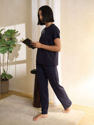 A woman in navy blue pajamas, featuring a white floral print on the top, stands reading a book.