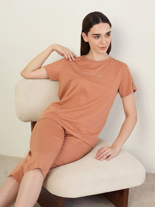 A woman in a terracotta-colored short-sleeve top and shorts set, with "your own story" printed on the top, sits casually on a chair.