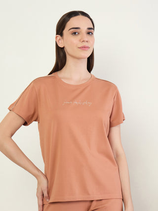 Women in a relaxed pose, wearing a peach-colored t-shirt with a subtle "your own story" print, featuring contrasting trim on the sleeves.