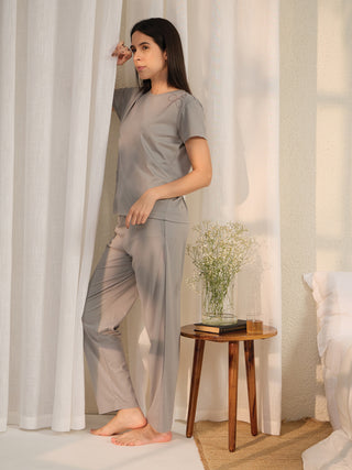A woman in a grey pajama set with subtle embroidery stands beside a white curtain, looking to the side.