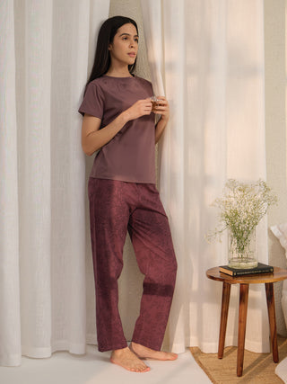 A woman in a mauve t-shirt with floral embroidery and mauve printed lounge pants holds a glass of tea while standing by a window.