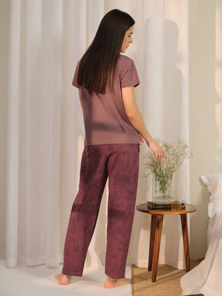 A woman stands with her back to the camera, wearing a mauve t-shirt and burgundy floral print pajama pants.