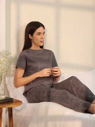 A woman in a charcoal grey pajama set with lace print pants and floral embroidery on the top, sits on a bed holding a cup of tea.