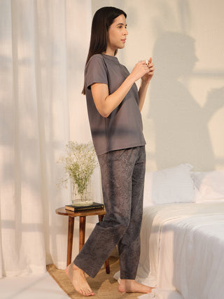 A woman stands in a grey pajama set with a dark grey floral print on the pants and subtle grey embroidery on the top.