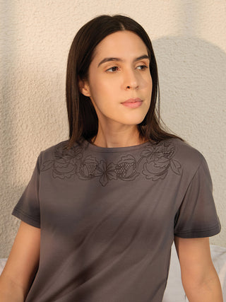 A woman is featured wearing a grey t-shirt with floral embroidery on the yoke and grey pajama pants.