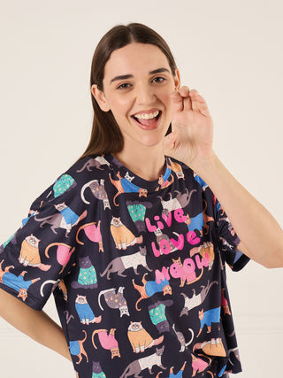 A woman in a dark blue t-shirt with a playful cat print, striking a cheerful pose with her hand near her mouth.