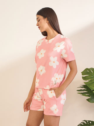 A woman stands facing left, wearing a pink floral-print pajama set with a scoop neck and short sleeves.