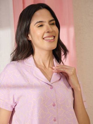 A woman in a lavender pajama top with a yellow floral print, smiling and resting her hand on her neck.