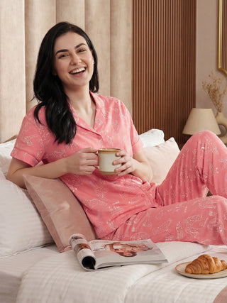Women in pink pajamas with a playful dog print lounging in bed with a mug and croissant.