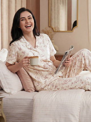 Woman in cream-colored pajamas with a playful red dog print, posing from the side, holding a mug and a magazine.
