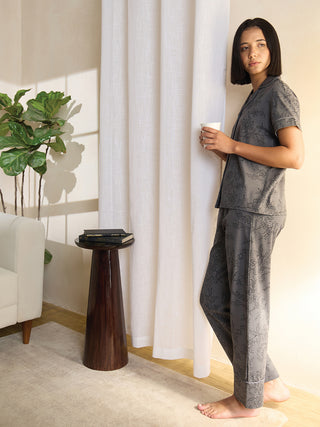 A woman in a gray floral print pajama set stands near a white curtain, holding a mug.