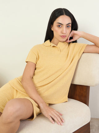 A woman in a yellow textured knit pajama set, featuring a collared short-sleeve top and matching shorts, poses confidently on a chair.