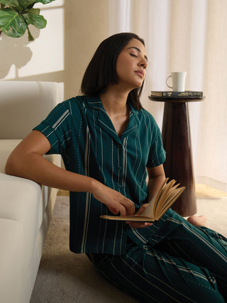 A woman in green striped sleepwear sits on the floor, reading a book with her eyes closed.
