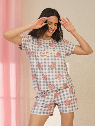 A woman poses happily in a light blue and white checkered pajama set featuring pink teddy bear prints and the word "Cozy" on the top.