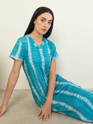 A woman in a teal tie-dye maxi dress is sitting on the floor, leaning against a white wall.