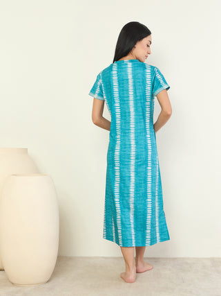 A woman stands facing away from the camera, wearing a teal and white tie-dye maxi dress with short sleeves.