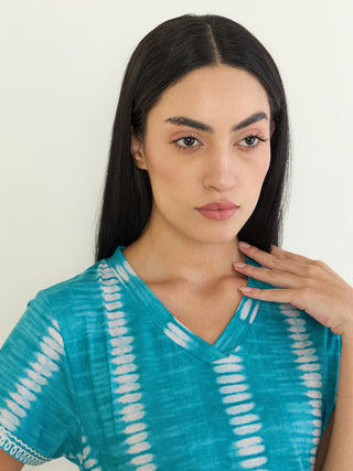 A woman stands with her shoulder facing forward, wearing a teal and white tie-dye V-neck t-shirt.