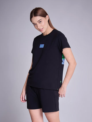 A woman poses, wearing a black t-shirt with a blue rectangular graphic on the left chest and a vertical abstract green and blue print on the right side, paired with black shorts.