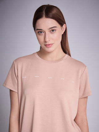 Woman standing in a front-facing close-up pose, wearing a warm taupe short-sleeve sleepwear T-shirt with minimal white word print across the chest.