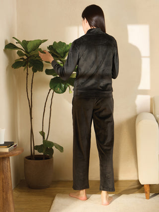 A woman in black velour pajamas with a zip-up jacket and loose-fitting pants stands facing away from the camera, tending to a large potted plant.