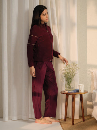 A woman in a burgundy velvet tracksuit, looking away from the camera, stands next to a small wooden table.