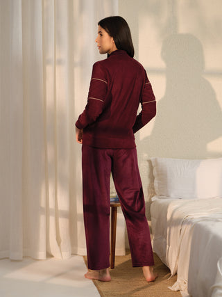 A woman stands facing away from the camera in a burgundy velour loungewear set, unlike the previous image, this one features a light-colored, textured wall in the background.
