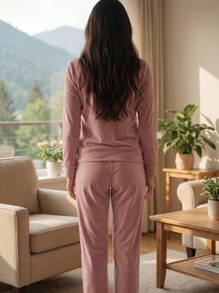 Women in a soft pink velvet pajama set, standing with their back to the camera, facing a window with a mountain view.