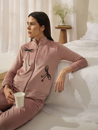 A woman in a pink velvet pajama set with black bow print, holding a white mug, poses seated on the floor next to a bed.